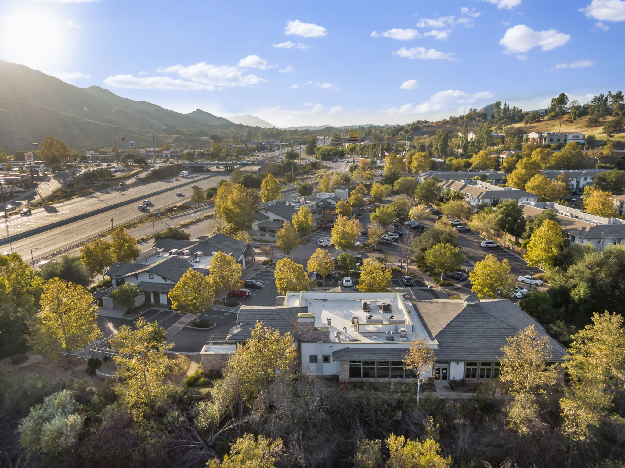 Golden hour aerial of The Shops at Oak Creek tucked into mature trees with the Santa Monica Mountains and 101 freeway visible behind